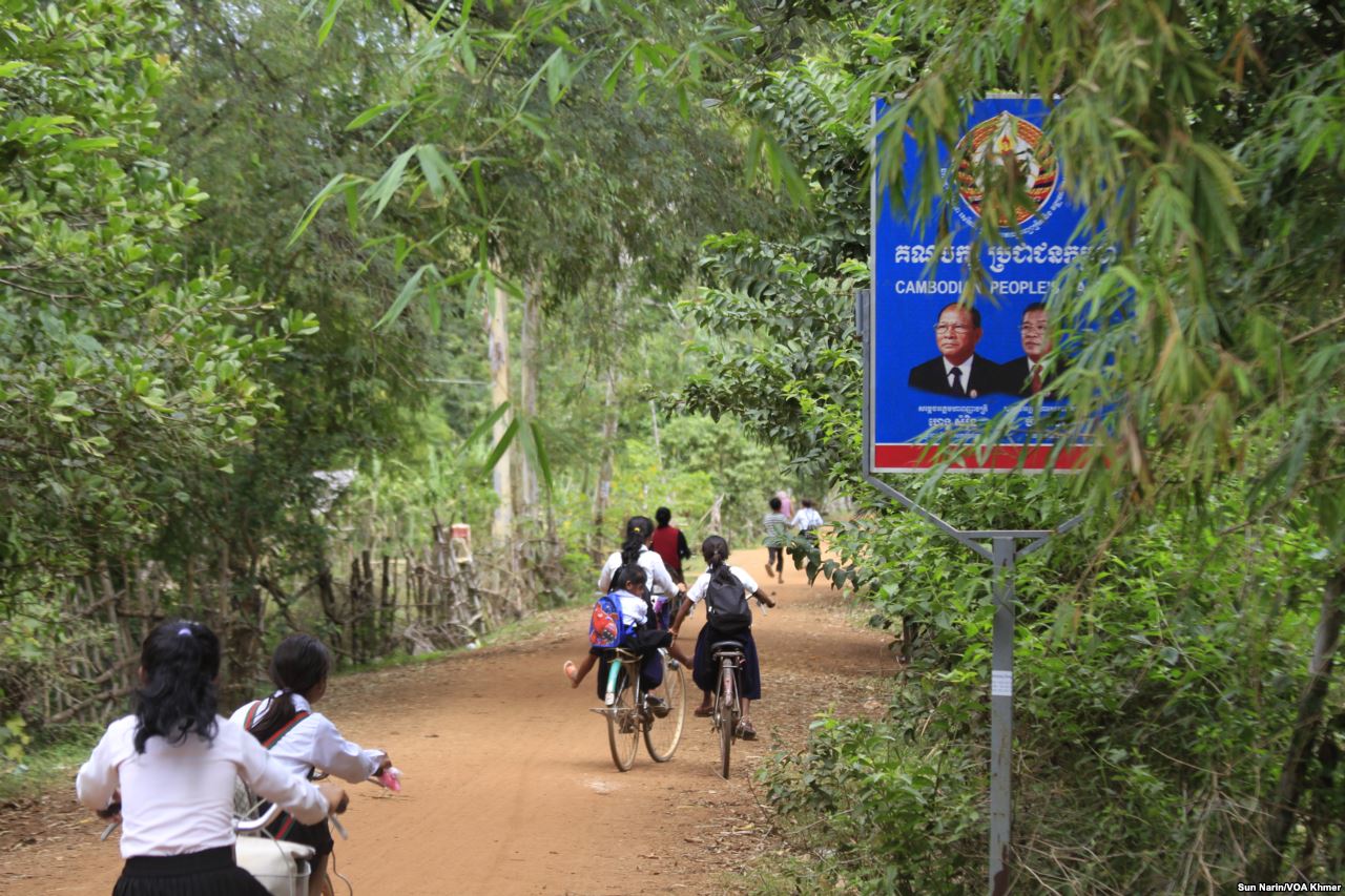 Children ride their bicycles past a sign bearing the Cambodian People's Party logo and the faces of Prime Minister Hun Sen and National Assembly President Heng Samrin